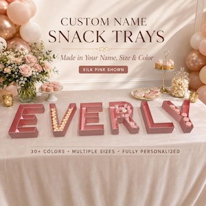 Puede incluir: Bandejas de aperitivos personalizadas en color rosa seda que deletrean EVERLY, rellenas de varios dulces y galletas. La imagen incluye el texto CUSTOM NAME SNACK TRAYS, Made in Your Name, Size & Color, SILK PINK SHOWN, y 30+ COLORS, MULTIPLE SIZES, FULLY PERSONALIZED.