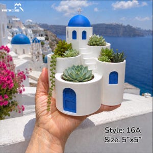 May include: A white planter shaped like a Greek church with a blue dome and doors, filled with succulents. The planter is 5 inches by 5 inches. The background shows a coastal town with similar architecture.
