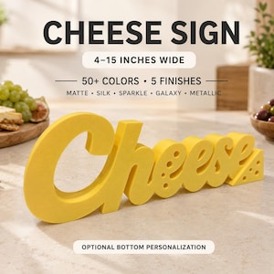 Pode incluir: Um letreiro 3D amarelo brilhante que soletra a palavra Cheese em fonte cursiva, com pequenos recortes circulares na última letra. O letreiro tem 10-38 cm de largura e está disponível em 50+ cores e 5 acabamentos: mate, seda, brilho, galáxia e metálico.