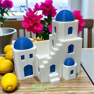 May include: A white 3D-printed model of a Greek-style building with blue domes and accents. The model has multiple levels, windows, and doors. The text "Style #1" is visible. The model is on a wooden surface with lemons.