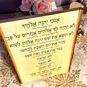 Op de afbeelding: Een houten plaquette met de Tien Geboden in het Hebreeuws. De tekst is in het hout gegraveerd en is duidelijk leesbaar. De plaquette is lichtbruin van kleur en heeft een natuurlijke houtnerf.