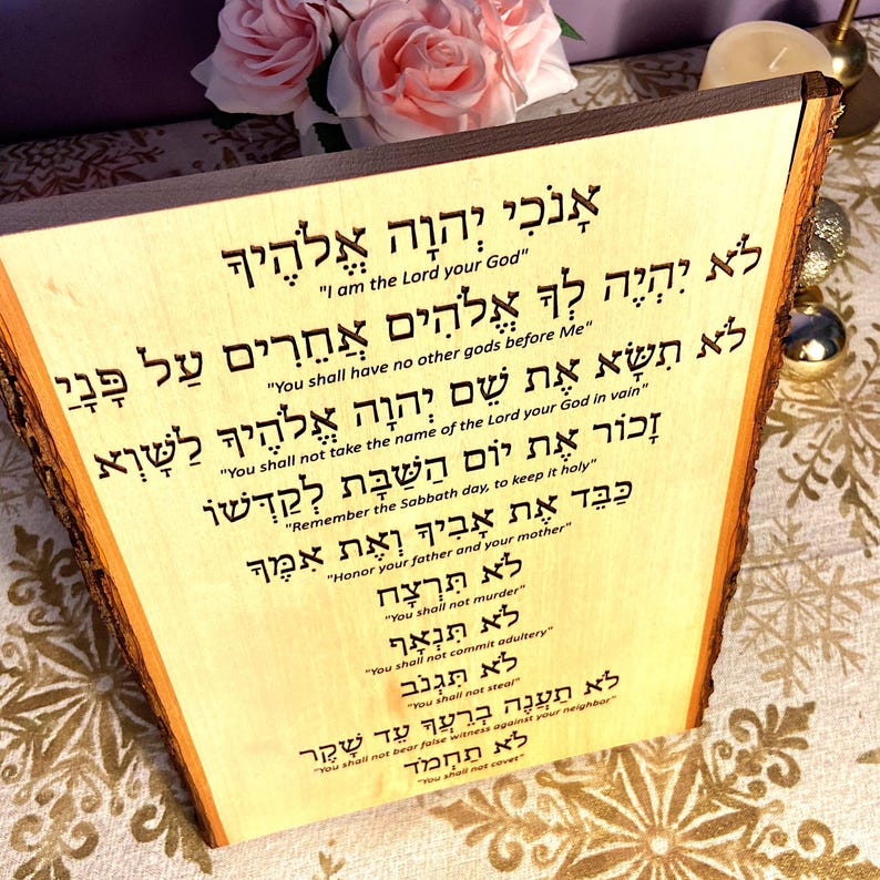 Op de afbeelding: Een houten plaquette met de Tien Geboden in het Hebreeuws. De tekst is in het hout gegraveerd en is duidelijk leesbaar. De plaquette is lichtbruin van kleur en heeft een natuurlijke houtnerf.