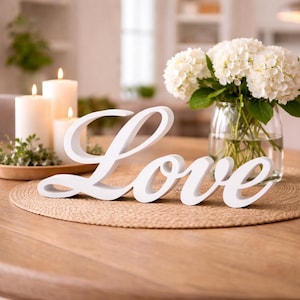 Può includere: Una scritta in legno bianco con la parola Love in carattere corsivo. L'oggetto è posizionato su una tovaglietta rotonda in paglia intrecciata color beige, accompagnato da un vaso in vetro trasparente con ortensie bianche e tre candele.