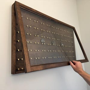 May include: A rectangular dark brown wooden display case with a hinged glass front door. The interior features a dark wood back panel with rows of small brass pegs for mounting items, with three circular silver coins displayed on the pegs.