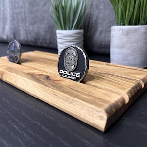 May include: A wooden display stand with three slots for displaying challenge coins. The stand is made of light brown wood and has a natural finish. A silver challenge coin with a black background and the words "POLICE CALGARY" is displayed in the centre slot.