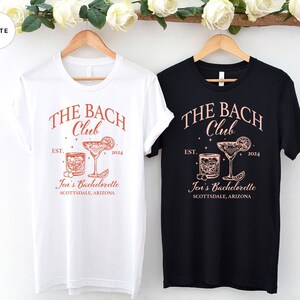 May include: Two t-shirts, one white and one black, with the text "THE BACH Club" in a red-orange color. The text "EST. 2024" and "Joi's Bachelorette Scottsdale, Arizona" are also printed on the shirts. The shirts feature a graphic of two cocktails.