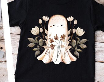 May include: Two t-shirts with a cute ghost design. The ghost is surrounded by flowers and leaves in shades of brown and white. The shirts are available in grey and black.