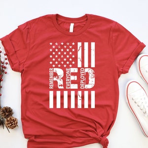 May include: Red t-shirt with a white distressed American flag design and the words "Remember Everyone Deployed Red" printed vertically on the flag.