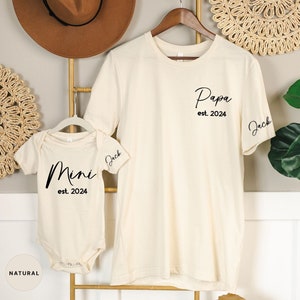 May include: A set of two cream-colored t-shirts with black text. The larger shirt says "Papa est. 2024" and has the name "Jack" on the sleeve. The smaller shirt says "Mini est. 2024" and also has the name "Jack" on the sleeve.