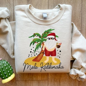 May include: A cream-colored sweatshirt with a graphic of a Santa Claus wearing sunglasses and holding a tropical drink. The Santa is standing in front of a palm tree with Christmas lights. The text "Mele Kalikimaka" is printed below the graphic.