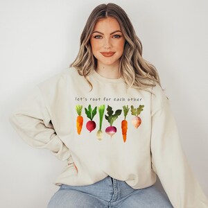 May include: A light beige sweatshirt with the text "Let's root for each other" and a graphic of colourful vegetables, including carrots, turnips, and radishes.