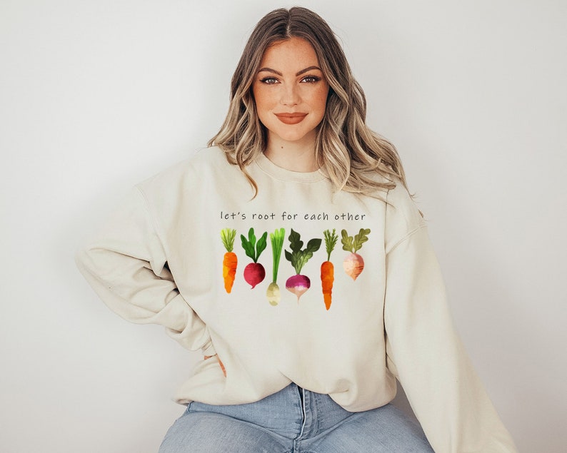 May include: A light beige sweatshirt with the text "Let's root for each other" and a graphic of colourful vegetables, including carrots, turnips, and radishes.