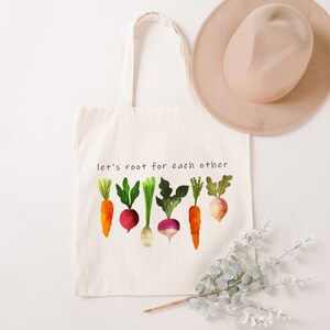 May include: A white canvas tote bag with the text "let's root for each other" printed on it. The bag features a colourful illustration of carrots, radishes, and turnips.