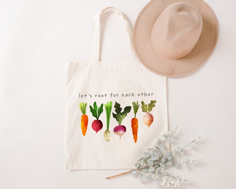 May include: A white canvas tote bag with the text "let's root for each other" printed on it. The bag features a colourful illustration of carrots, radishes, and turnips.