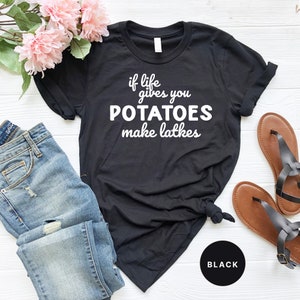 Peut inclure: T-shirt noir avec un texte blanc qui dit "if life gives you potatoes make latkes". Le t-shirt est pos&eacute; &agrave; plat sur une surface blanche avec un jean bleu et des sandales marrons.