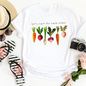 May include: White t-shirt with the text "Let's root for each other" and a watercolour illustration of carrots, radishes, and turnips.