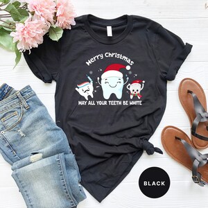 May include: Black t-shirt with a Christmas-themed dental graphic. The design features three cartoon teeth wearing Santa hats and holding toothbrushes. The text reads "Merry Christmas, may all your teeth be white."