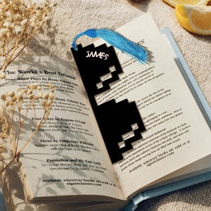 Op de afbeelding: Een zwarte boekenlegger met de naam "JAMES" in het wit, in een open boek. De boekenlegger heeft een gepixeld ontwerp en een blauwe kwast. Het boek heet "The World's Best Drama."