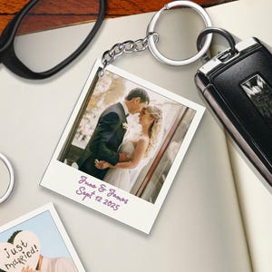 May include: A silver keychain with a photo of a couple on their wedding day. The photo is in a polaroid style with the text "June & James Sept 12 2025". A black car key fob is next to the keychain.