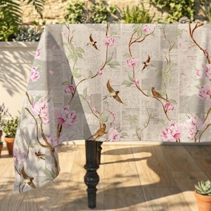Könnte beinhalten: Rechteckige Tischdecke mit Blumen- und Vogelmuster. Das Design zeigt rosa Pfingstrosen, braune Zweige und kleine Vögel auf einem hellgrauen Hintergrund mit Zeitungsmuster. Die Tischdecke liegt auf einem dunklen Holztisch.