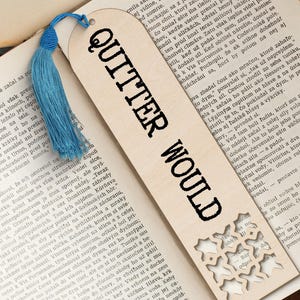 Op de afbeelding: Een houten bladwijzer met de tekst "QUITTER WOULD" in zwarte letters. Een blauwe kwast is aan de bovenkant bevestigd. De bladwijzer ligt op de open pagina's van een boek.
