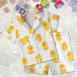 May include: White pajama set with a repeating pattern of yellow cartoon ducklings. The set includes a button-down shirt with a pocket and matching shorts. The pajamas are designed for children.