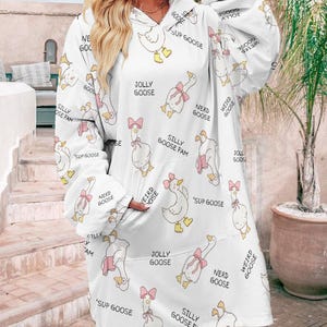 Op de afbeelding: Witte hoodie met een grillig ganzenpatroon. Het ontwerp toont cartoon ganzen met roze strikken en teksten als "Jolly Goose" en "Nerd Goose". De hoodie heeft een grote voorzak en lange mouwen.