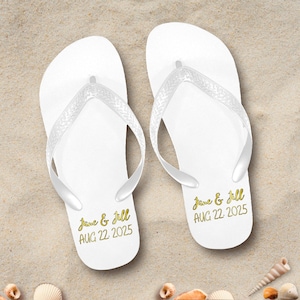May include: A pair of white flip-flops with clear straps, personalized with gold text reading "Jane & Jill AUG 22. 2025". The sandals are resting on a sandy surface, suggesting a beach or vacation theme.