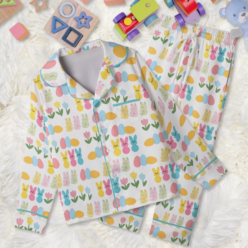 May include: White pajama set with a colorful Easter-themed pattern. The set includes a button-down shirt and pants, both adorned with bunnies, eggs, and flowers in pastel colors. The pajamas have light blue trim.