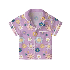 Puede incluir: Camiseta de pijama de manga corta lila, adornada con un estampado floral repetido en tonos amarillos, azules, rosas y blancos. La camisa presenta un cuello cl&aacute;sico, un bolsillo en el pecho y ribetes blancos en los bordes.