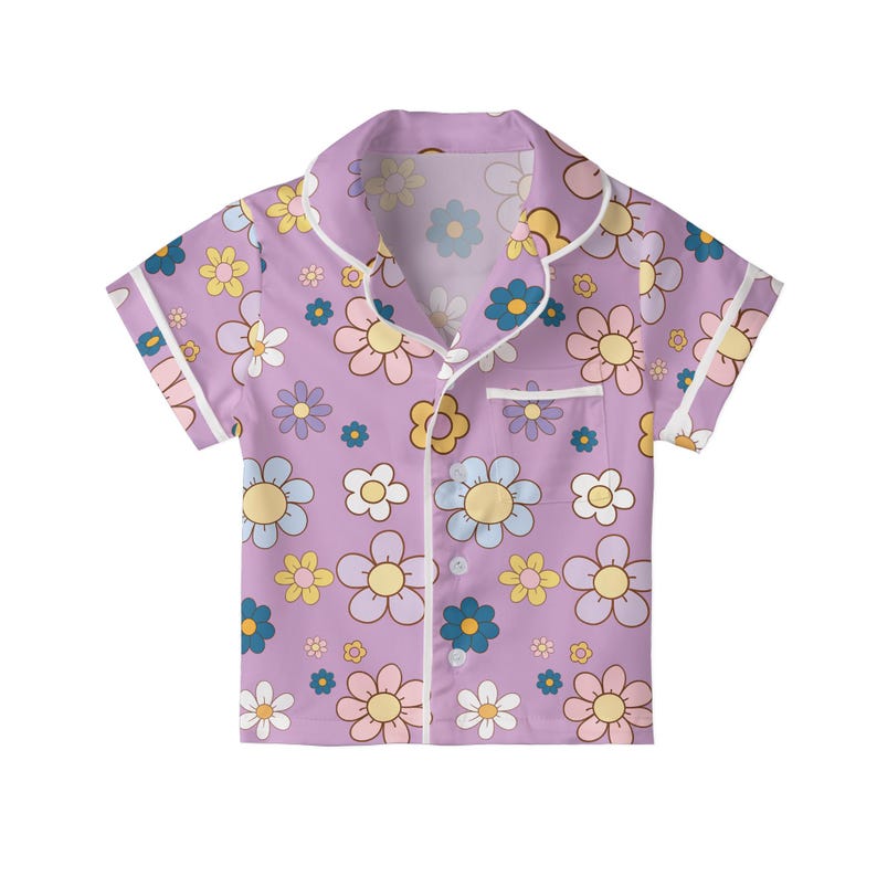 Puede incluir: Camiseta de pijama de manga corta lila, adornada con un estampado floral repetido en tonos amarillos, azules, rosas y blancos. La camisa presenta un cuello cl&aacute;sico, un bolsillo en el pecho y ribetes blancos en los bordes.