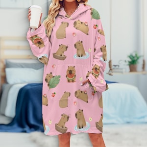 May include: A pink hooded blanket with a repeating pattern of cartoon capybaras, watermelon slices, and other fruits. The blanket has a large front pocket and long sleeves. The capybaras are in various poses, some in water, others holding watermelon.