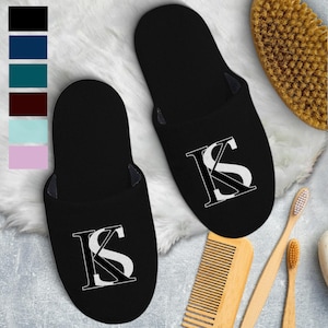 May include: Black house slippers with a white "KS" monogram. The slippers are displayed on a white, fluffy surface, with a color palette of black, navy, teal, burgundy, light blue, and lavender. Other bath accessories are also visible.