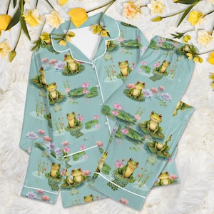 May include: Light blue pajama set featuring a whimsical frog pond design. The set includes a button-down top and matching pants, both adorned with illustrations of frogs on lily pads, pink water lilies, and white trim.