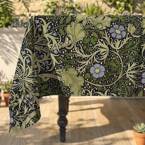 May include: A rectangular tablecloth featuring a dense botanical pattern with light green leaves, dark green foliage, and pale blue flowers set against a dark charcoal background. The fabric drapes over the edge of a table with a visible corner fold.