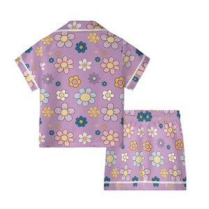 Puede incluir: Conjunto de pijama lila con una camisa de manga corta y pantalones cortos a juego. El conjunto presenta un estampado repetido de flores retro de colores en tonos amarillos, rosas, azules y blancos. Los bordes est&aacute;n ribeteados con ribetes blancos.