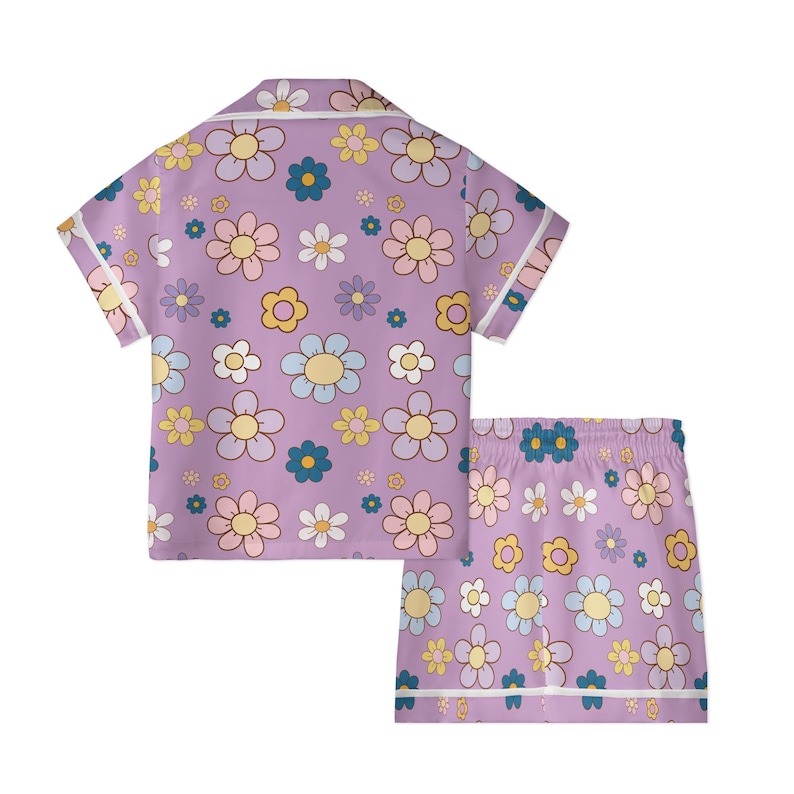 Puede incluir: Conjunto de pijama lila con una camisa de manga corta y pantalones cortos a juego. El conjunto presenta un estampado repetido de flores retro de colores en tonos amarillos, rosas, azules y blancos. Los bordes est&aacute;n ribeteados con ribetes blancos.