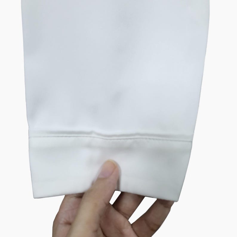 May include: Close-up of a white fabric cuff with a stitched hem. The fabric appears smooth and opaque, with a slight sheen. The cuff is held by a hand, suggesting it is part of a garment.