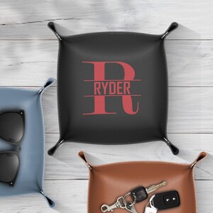 Op de afbeelding: Een zwart leren valet tray met de naam "Ryder" in rode letters. De tray is vierkant met afgeronde hoeken en opstaande randen. Andere trays in blauw en bruin zijn ook zichtbaar.