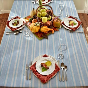 May include: A festive table setting with a light blue and white striped tablecloth. The table is set with white plates, red napkins, silverware, and wine glasses. A centerpiece includes a basket of apples, pumpkins, gourds, and fall foliage.