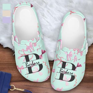 May include: A pair of light teal clogs with a white interior, decorated with pink bows and ribbons. Each clog features the name "Barbara" beneath a large black letter "D". The clogs are personalized and suitable for casual wear.