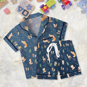 Puede incluir: Conjunto de pijama azul con estampado de animales del bosque, incluyendo zorros, ciervos y osos. El conjunto presenta una camisa de manga corta con botones y pantalones cortos a juego con cord&oacute;n blanco. Ribete negro.