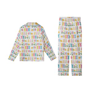 May include: White pajama set with a colorful Easter-themed pattern. The set includes a long-sleeved top and pants, both adorned with bunnies, eggs, and flowers in pastel colors. The trim is a light blue.