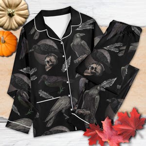 Pode incluir: Conjunto de pijama preto com um padrão repetido de corvos, caveiras e penas. A parte superior de manga comprida e as calças têm debrum branco e fechos de botões. O design é sobre um fundo preto.