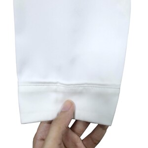 Puede incluir: Primer plano de un pu&ntilde;o de tela blanca con dobladillo cosido. La tela parece lisa y opaca. El pu&ntilde;o est&aacute; sujeto por una mano, con los dedos visibles en la parte inferior de la imagen. El fondo es blanco liso.