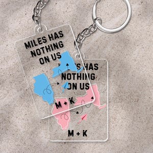 Könnte beinhalten: Zwei transparente Acryl-Schlüsselanhänger mit silbernen Ketten und Schlüsselringen. Jeder Schlüsselanhänger hat eine rechteckige Form mit dem Text "MILES HAS NOTHING ON US" und einem Bundesstaaten-Kartendesign. Eine Karte ist blau, die andere rosa, mit den Initialen "M + K".