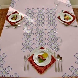 Peut inclure: Une nappe rose pâle ornée d'une bande centrale verticale de motifs floraux géométriques bleus et jaunes, avec des motifs assortis aux coins. Trois assiettes blanches sont posées sur des sets de table à franges rouille avec couverts et verres.