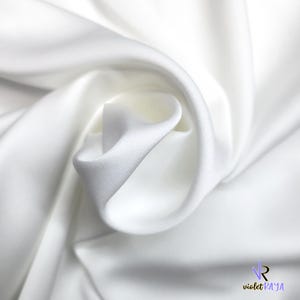 Puede incluir: Primer plano de tela blanca con pliegues suaves y textura sedosa. La tela est&aacute; drapeada, creando una sensaci&oacute;n de movimiento y profundidad. La imagen resalta la naturaleza suave y fluida del material, ideal para ropa o decoraci&oacute;n del hogar.