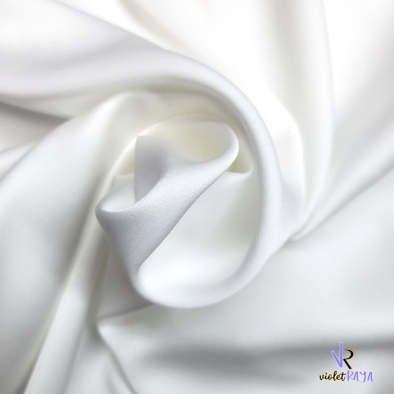 Puede incluir: Primer plano de tela blanca con pliegues suaves y textura sedosa. La tela est&aacute; drapeada, creando una sensaci&oacute;n de movimiento y profundidad. La imagen resalta la naturaleza suave y fluida del material, ideal para ropa o decoraci&oacute;n del hogar.