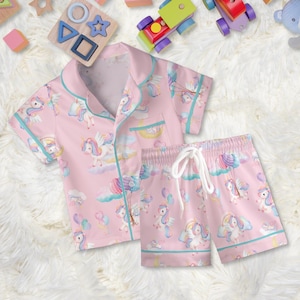 May include: Pink pajama set featuring a unicorn print with clouds, rainbows, and balloons. The set includes a short-sleeved button-up shirt and matching shorts with a drawstring waist. The trim is a light teal color.